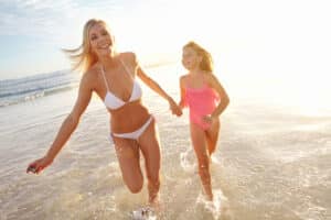 blonde mother in white bikini holding the hand of her blond daughter running down a beach at sunset