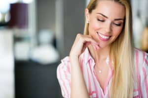 blonde female in pink and white shirt smiling and looking down with her hand below her chin